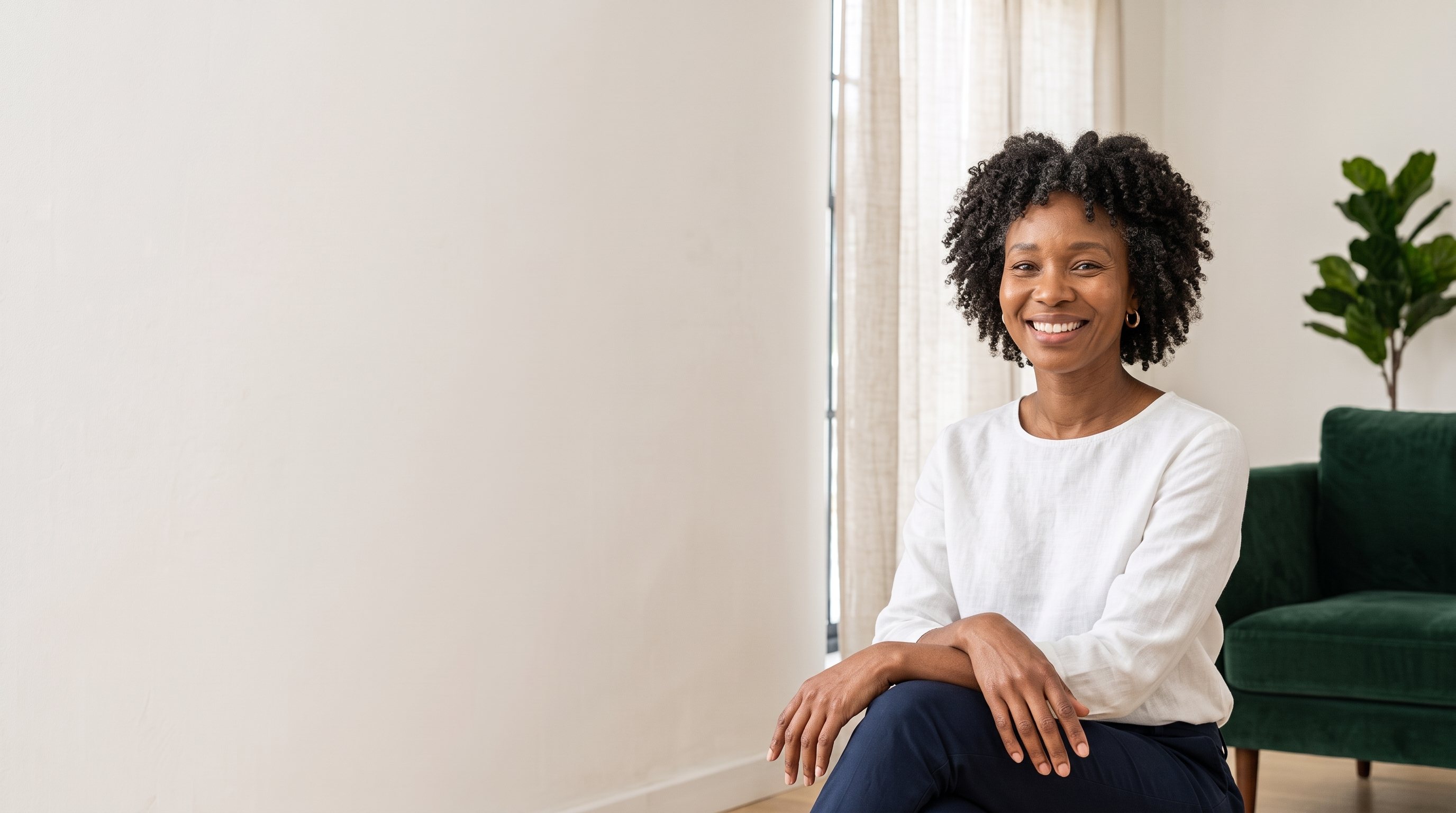 A smiling woman in a white linen top sitting in a bright living room beside a green armchair